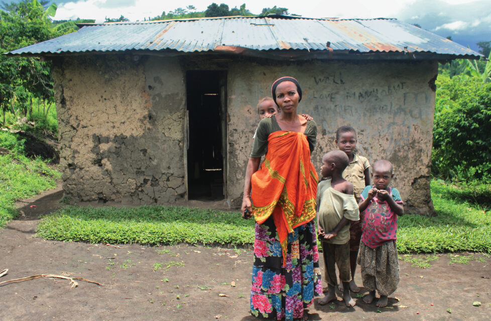 Ugandan Widow Faithful to Christ, Loses Second Husband A woman standing outside in front of her house with her four children